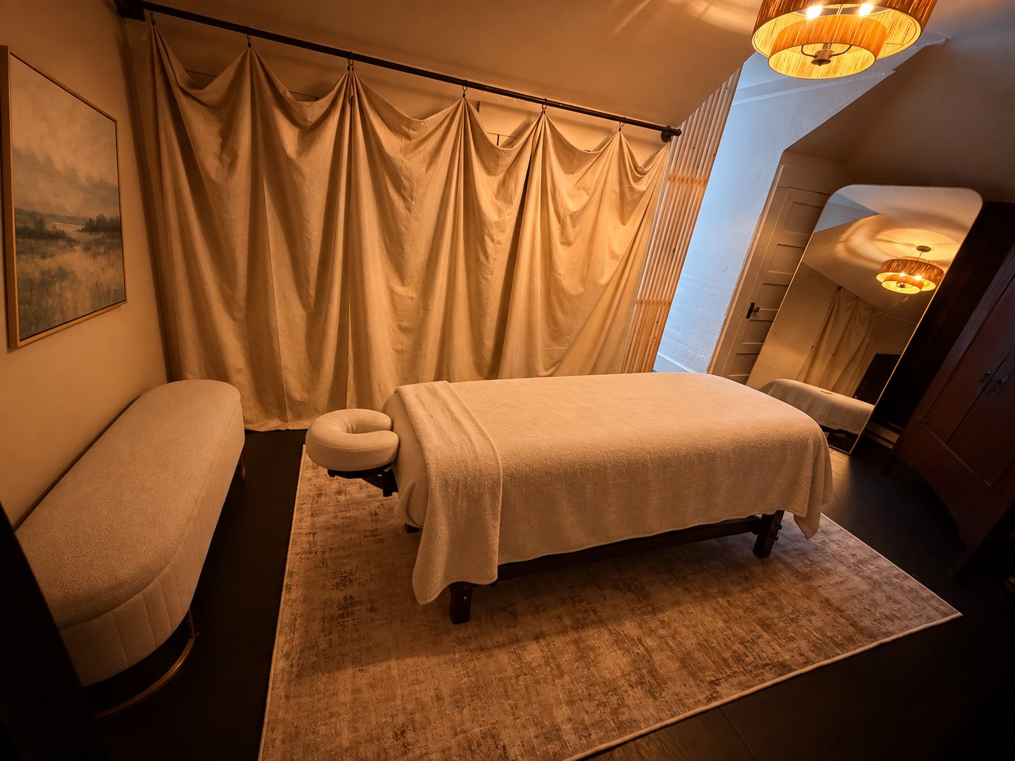 A softly lit treatment room with a made-up massage table, linen drapery, and a plush bench.